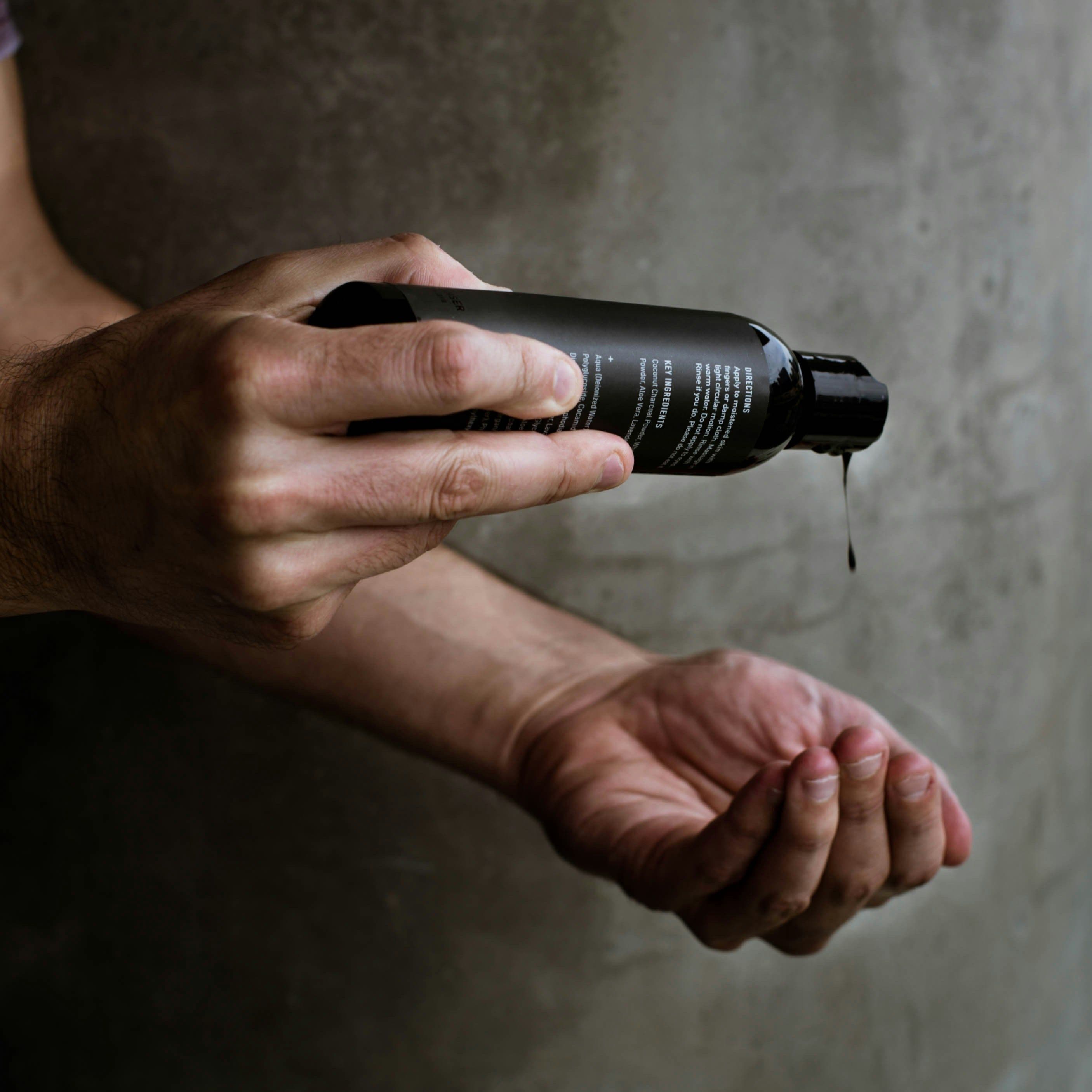 Close-up of a man’s hands pouring a dark liquid body wash from a sleek black bottle against a minimalist concrete background, emphasizing masculinity and premium skincare.